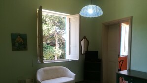 Una habitación con un sillón blanco, una mesa, suelo estampado, una ventana con vista a árboles y una lámpara colgante.