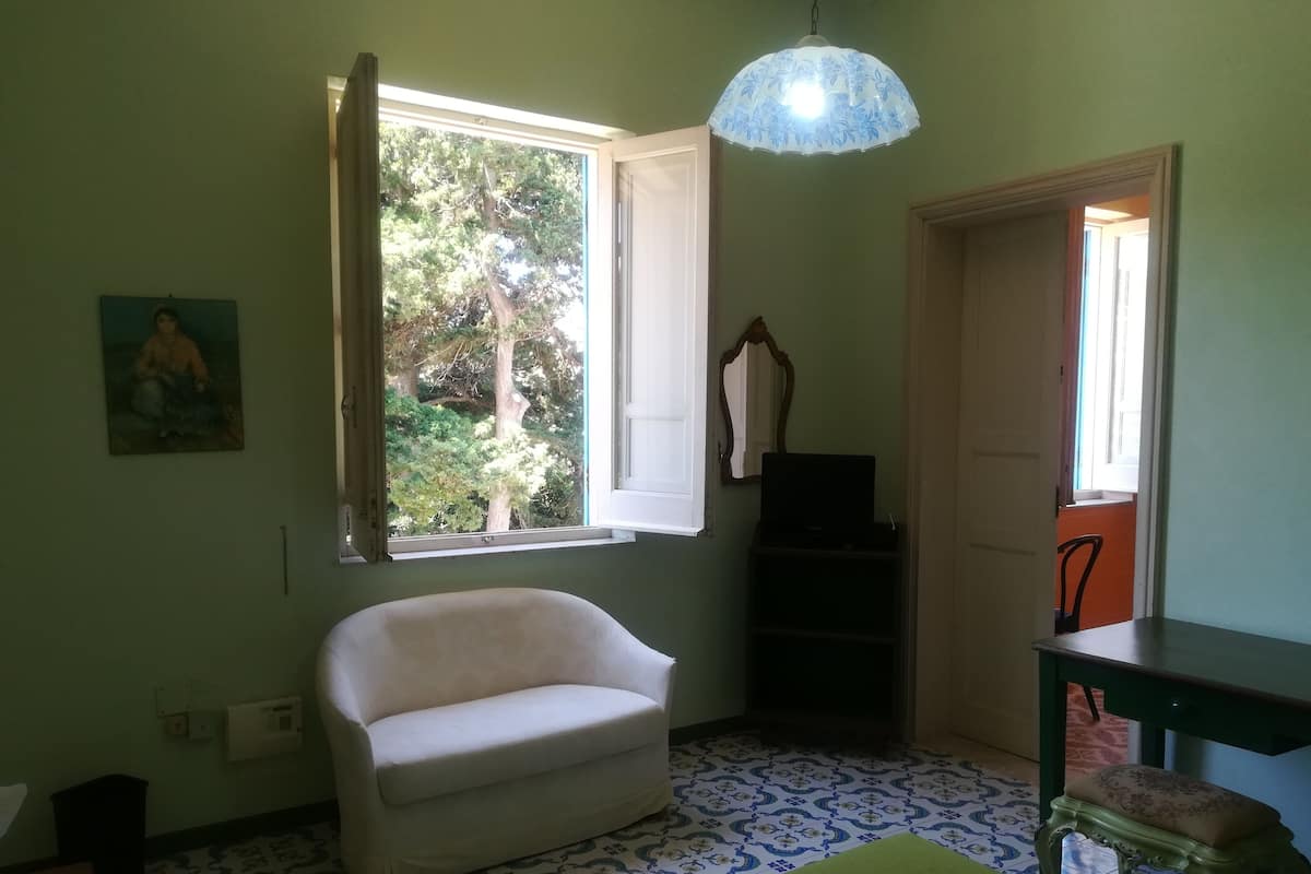 A room with a white armchair, a table, a patterned floor, a window with a view of trees, and a hanging light fixture.