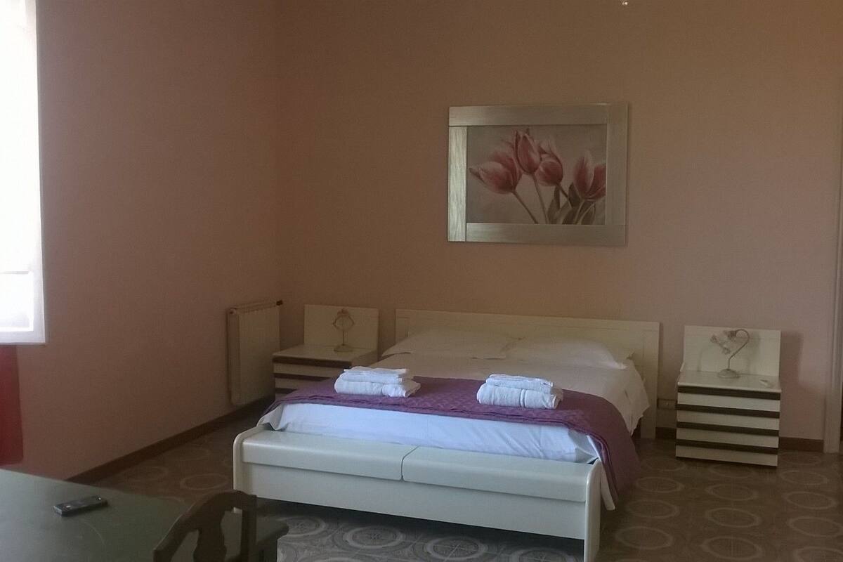 A bedroom with a bed, two bedside tables, a nightstand, a mirror, and a painting of flowers.