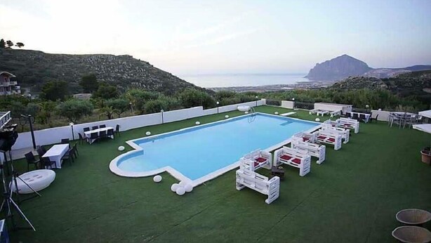 Una piscina en la azotea con sillones y mesas blancas, rodeada de césped artificial verde, con vista al mar y colinas al fondo.