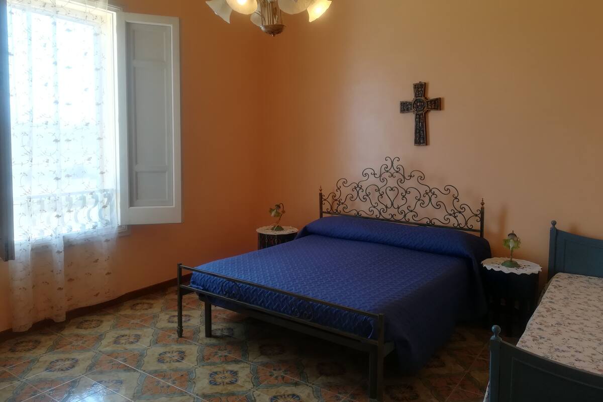 A bedroom with a bed, a cross on the wall, a small table, and a window with curtains.