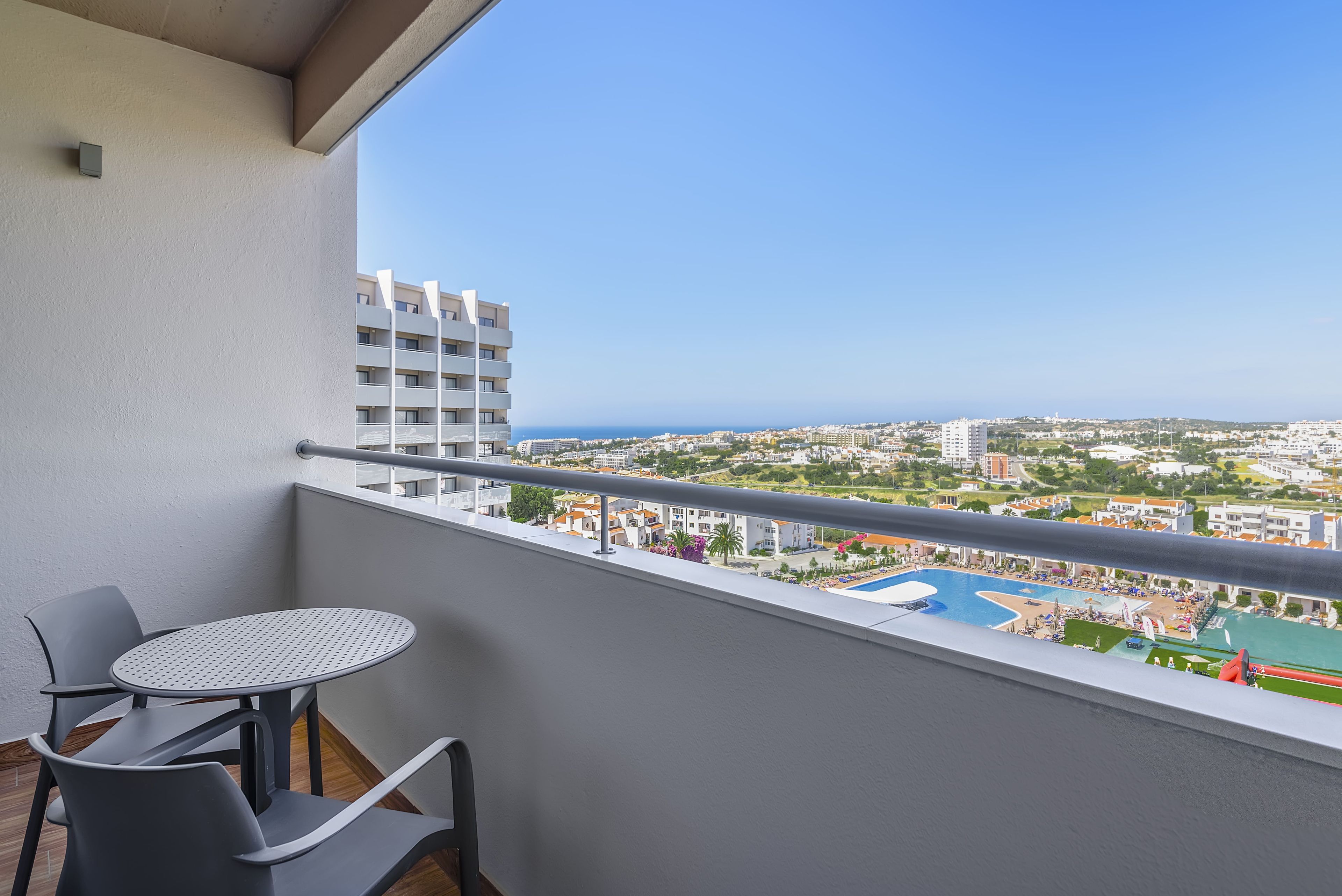 A balcony with a table and chairs, overlooking a cityscape and a swimming pool.