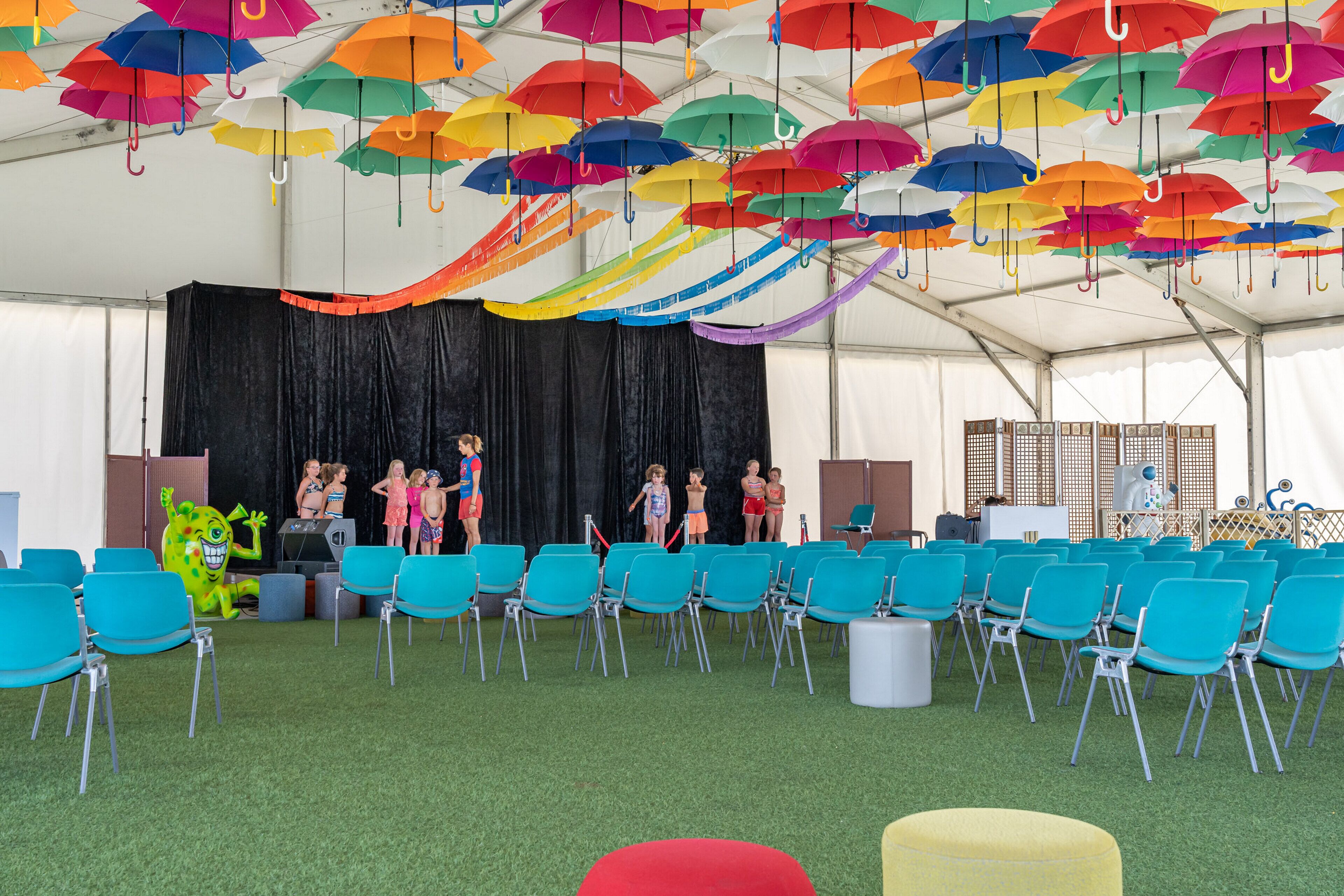A tent with colorful umbrellas hanging from the ceiling, teal chairs arranged in rows, and a stage with people.
