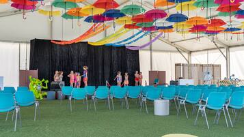 A tent with colorful umbrellas hanging from the ceiling, teal chairs arranged in rows, and a stage with people.