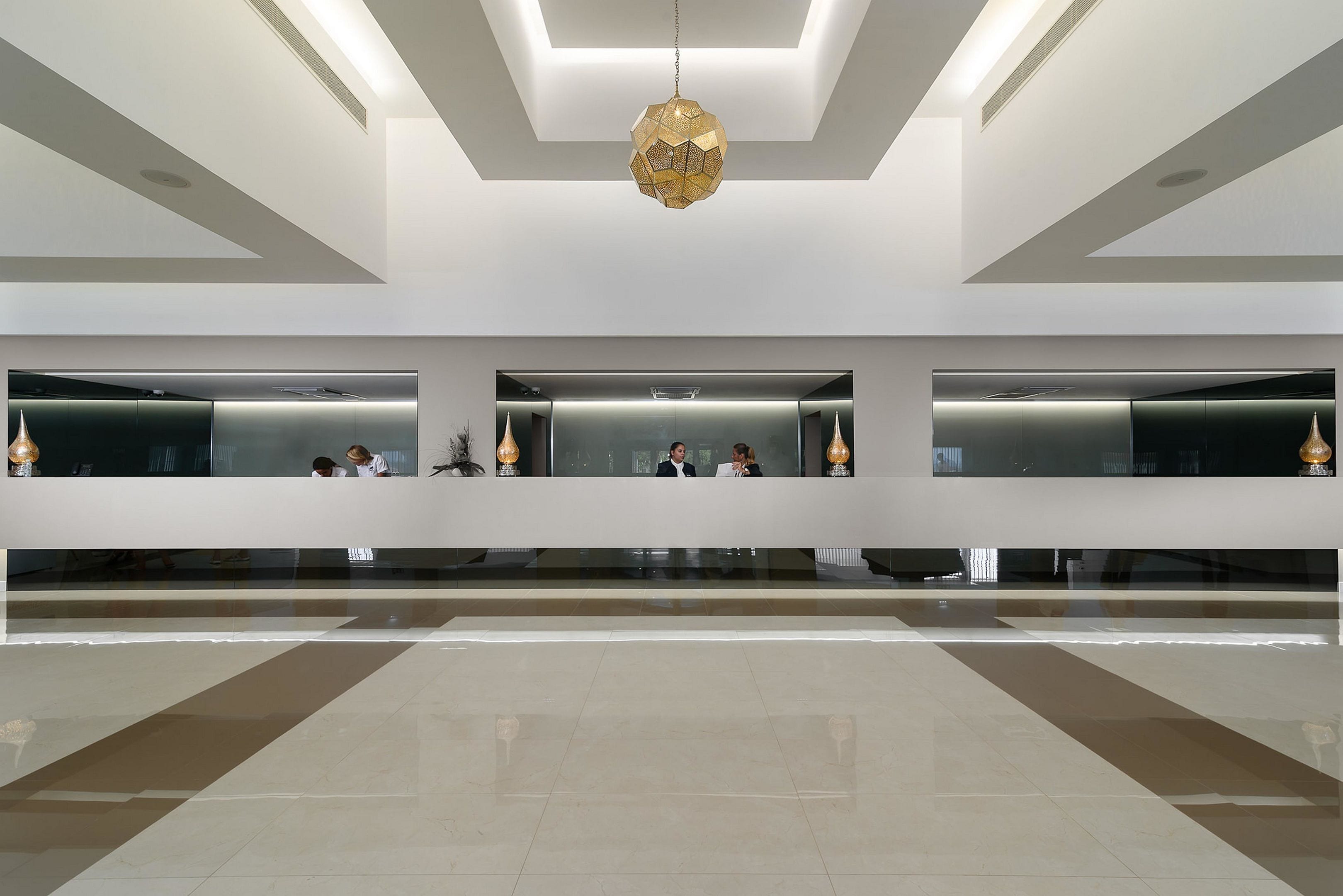 A spacious lobby with a modern design, featuring a central seating area with a geometric chandelier, a reception desk, and a clean, reflective floor.