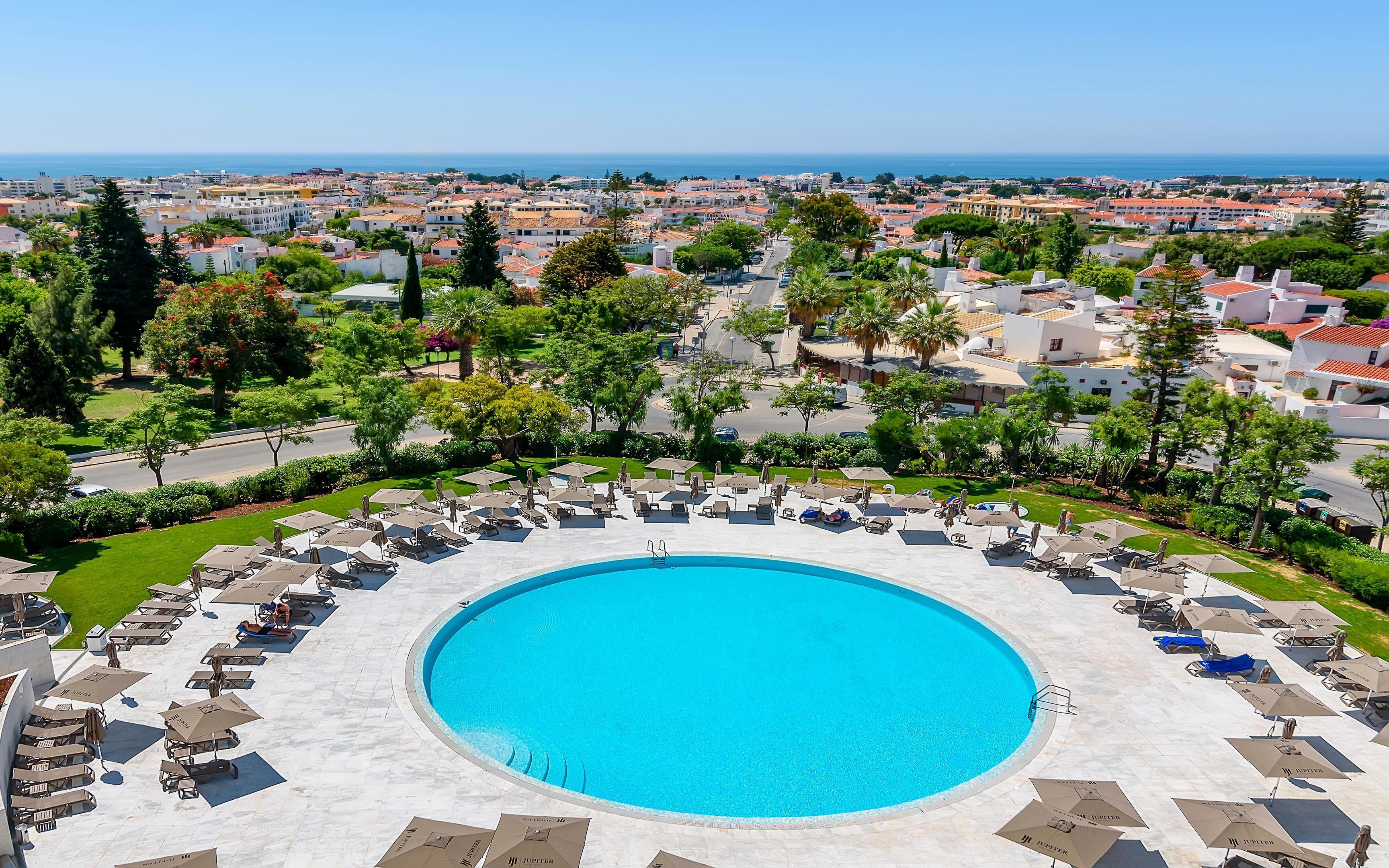 Une grande piscine ronde, entourée de chaises longues et de parasols, d’où l’on peut admirer une ville côtière en arrière-plan.