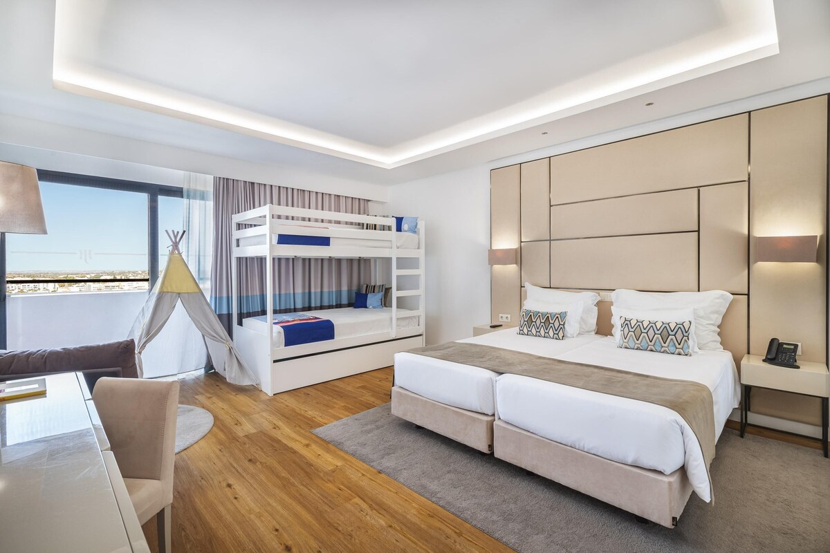 A modern hotel room with a bunk bed, a desk with a chair, a large window with a view, and a bedside table with a lamp.