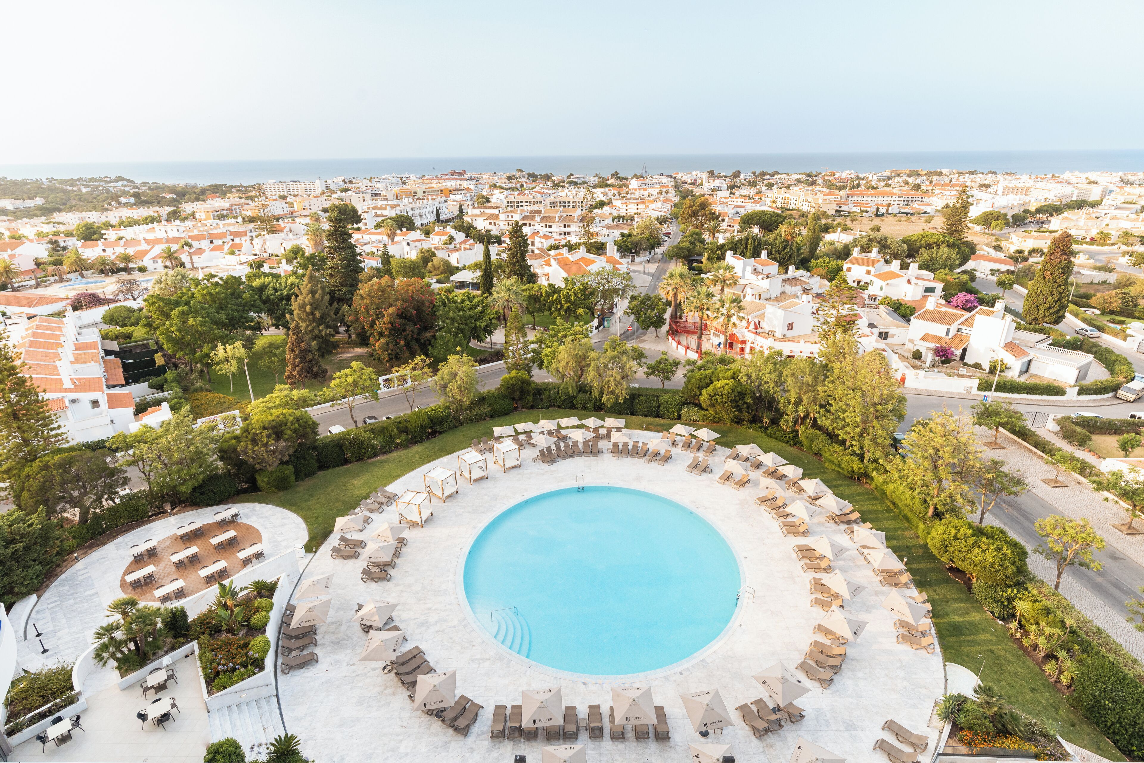 A large circular pool surrounded by lounge chairs and tables, with a view of a coastal town in the background.