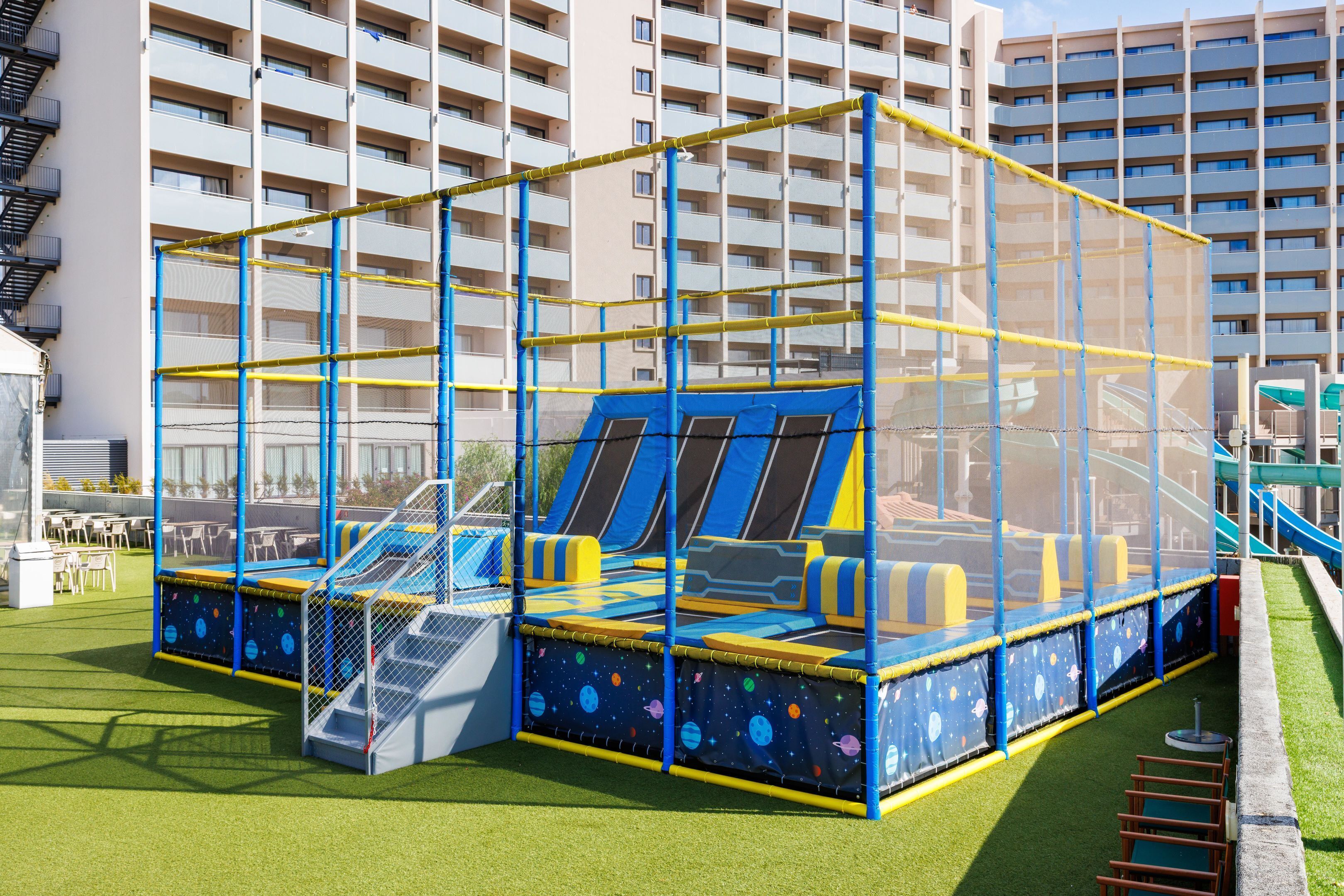 A children's play area with a trampoline and slides, surrounded by a safety net, in front of a multi-story hotel building.