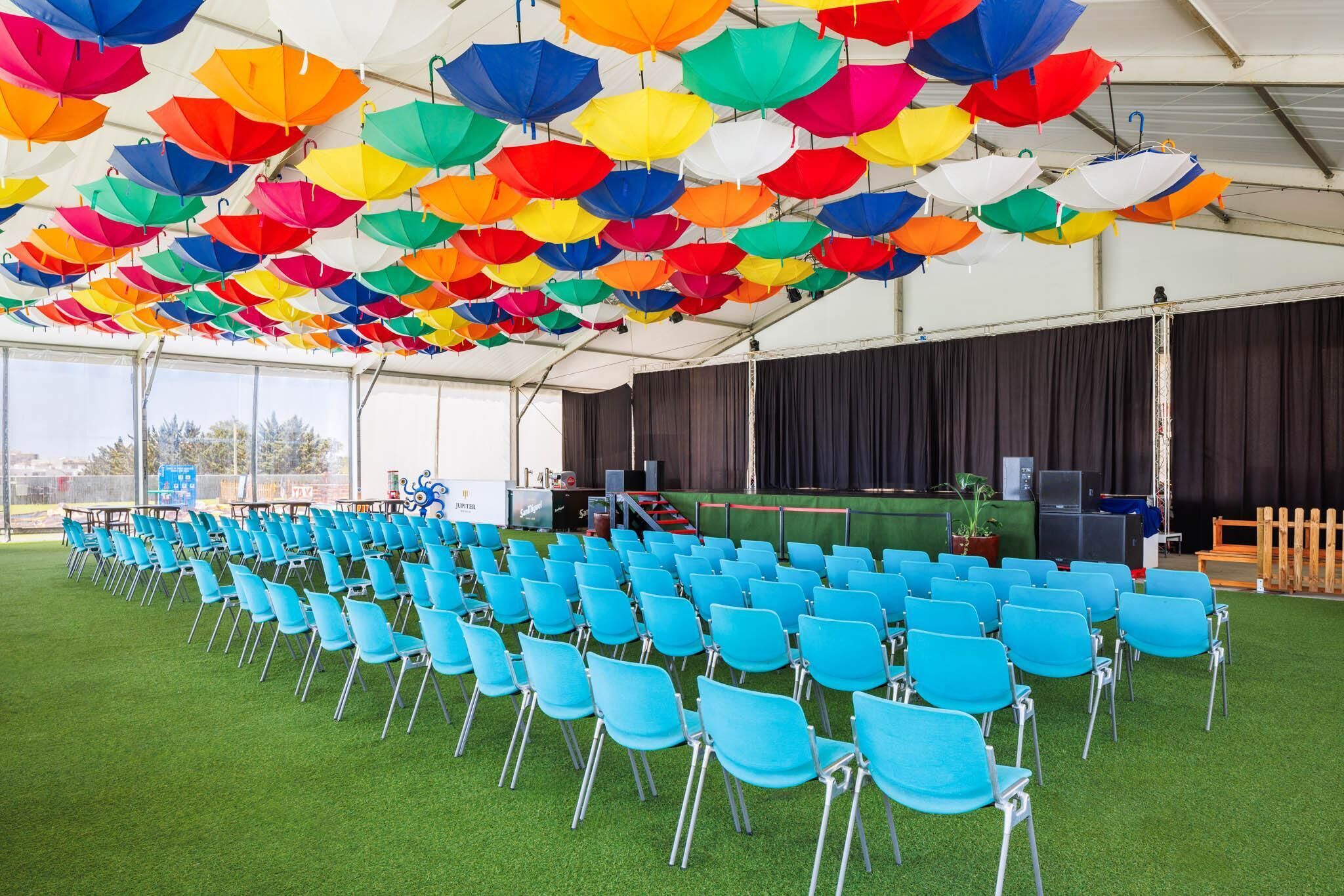 Une tente sous laquelle se trouvent des rangées de chaises bleues et des parasols colorés.