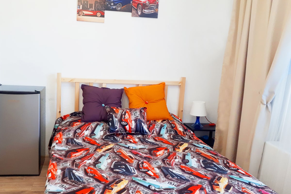 A bed with a car-themed bedspread, a small bedside table with a lamp, and a wall with three car posters.