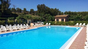 Una piscina rectangular con agua azul cristalina, rodeada de sillones y sombrillas blancas, con un edificio y árboles al fondo.