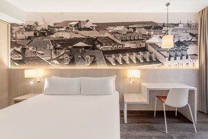A hotel room with a bed, a desk, a chair, and a cityscape mural on the wall.