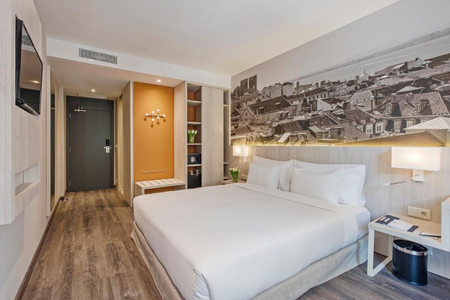 A hotel room with a large bed, a wooden floor, a wardrobe, a desk with a lamp, and a wall-mounted TV.