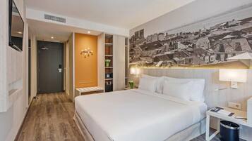 A hotel room with a large bed, a wooden floor, a wardrobe, a desk with a lamp, and a wall-mounted TV.