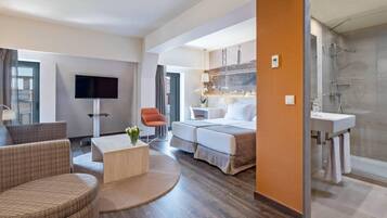 Une chambre d’hôtel moderne équipée d’un téléviseur à écran plat, d’un canapé, d’une table basse, d’un lit avec deux oreillers, d’une petite table avec une plante et d’un espace douche.