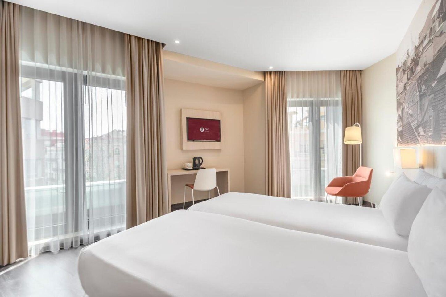 Habitación de hotel con una cama grande, un escritorio con silla, cafetera y vistas a la ciudad a través de amplios ventanales.