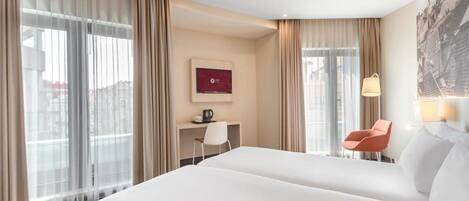 A hotel room with a large bed, a desk with a chair, a coffee maker, and a view of the city through large windows.