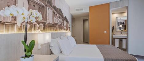 Habitación de hotel con cama, escritorio, orquídea en maceta, lámpara de pared, baño con espejo y un cuadro de un edificio.