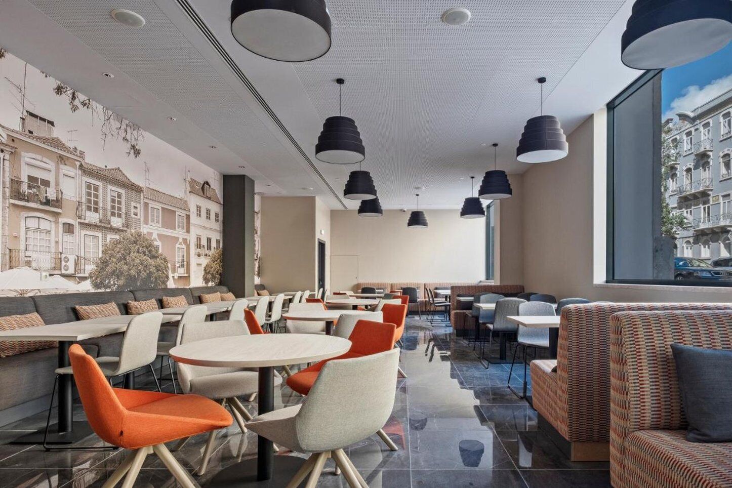 A modern restaurant with round tables and orange chairs, large black pendant lights, and a view of a historic building through large windows.