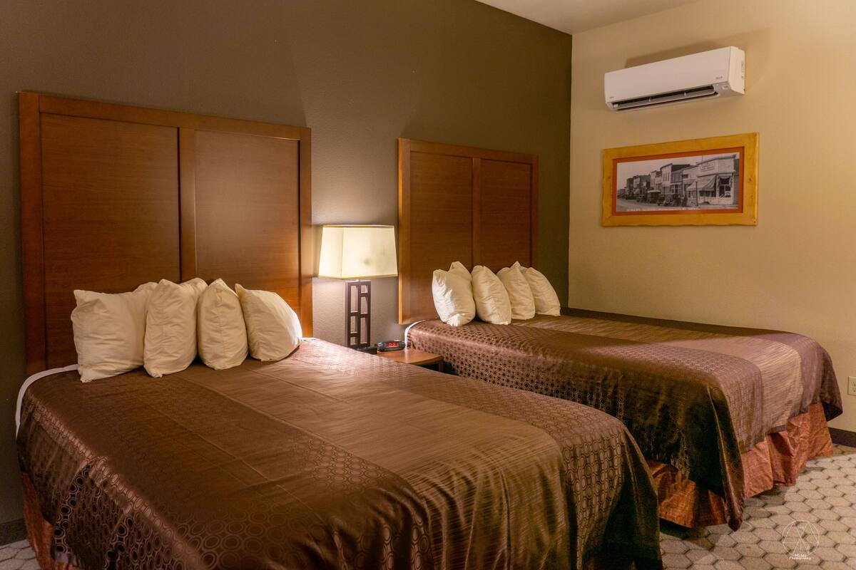 A hotel room with two beds, a nightstand, a lamp, an air conditioner, and a framed picture on the wall.