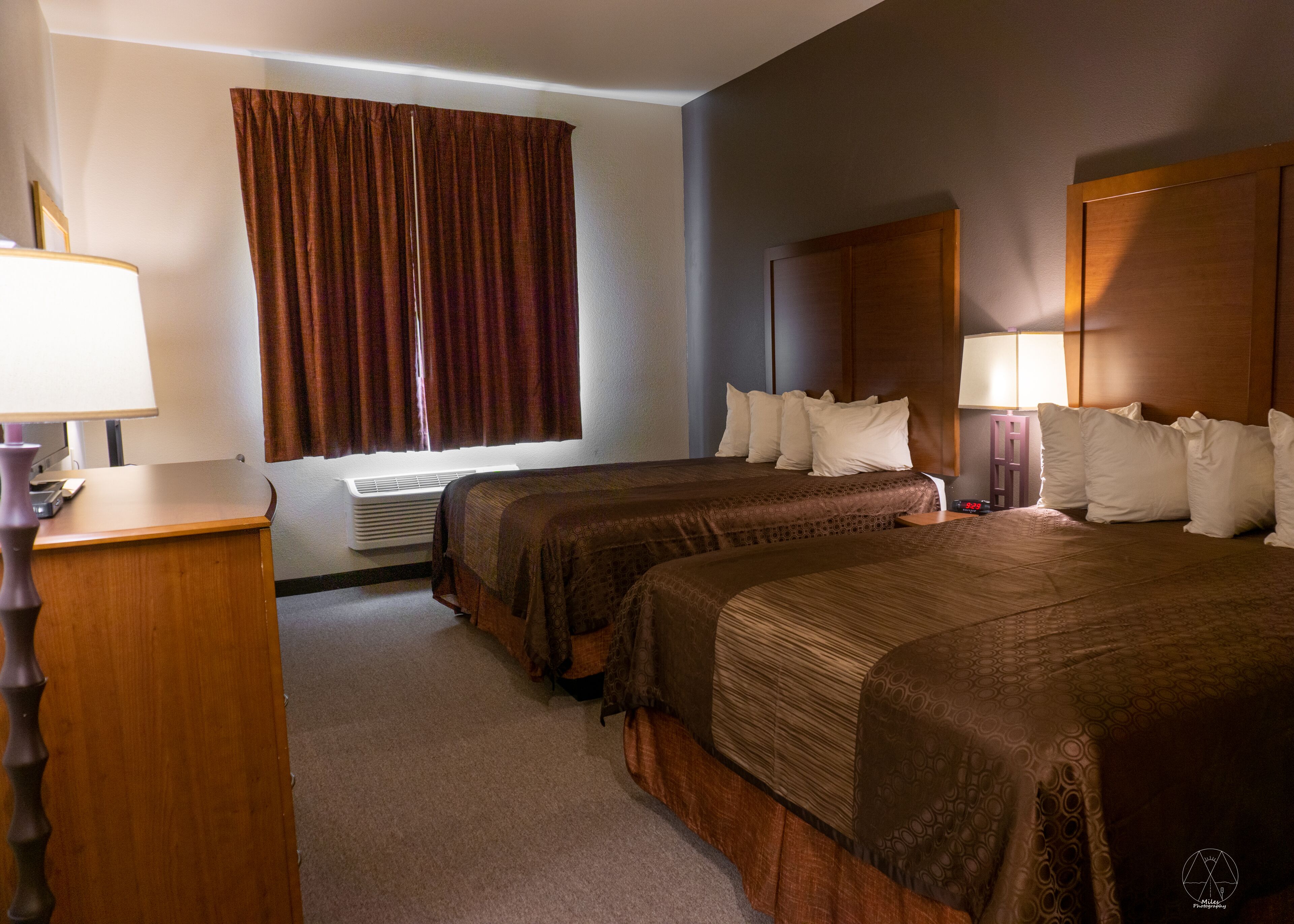 Quarto de hotel com duas camas, uma escrivaninha, uma luminária e uma janela com cortinas.