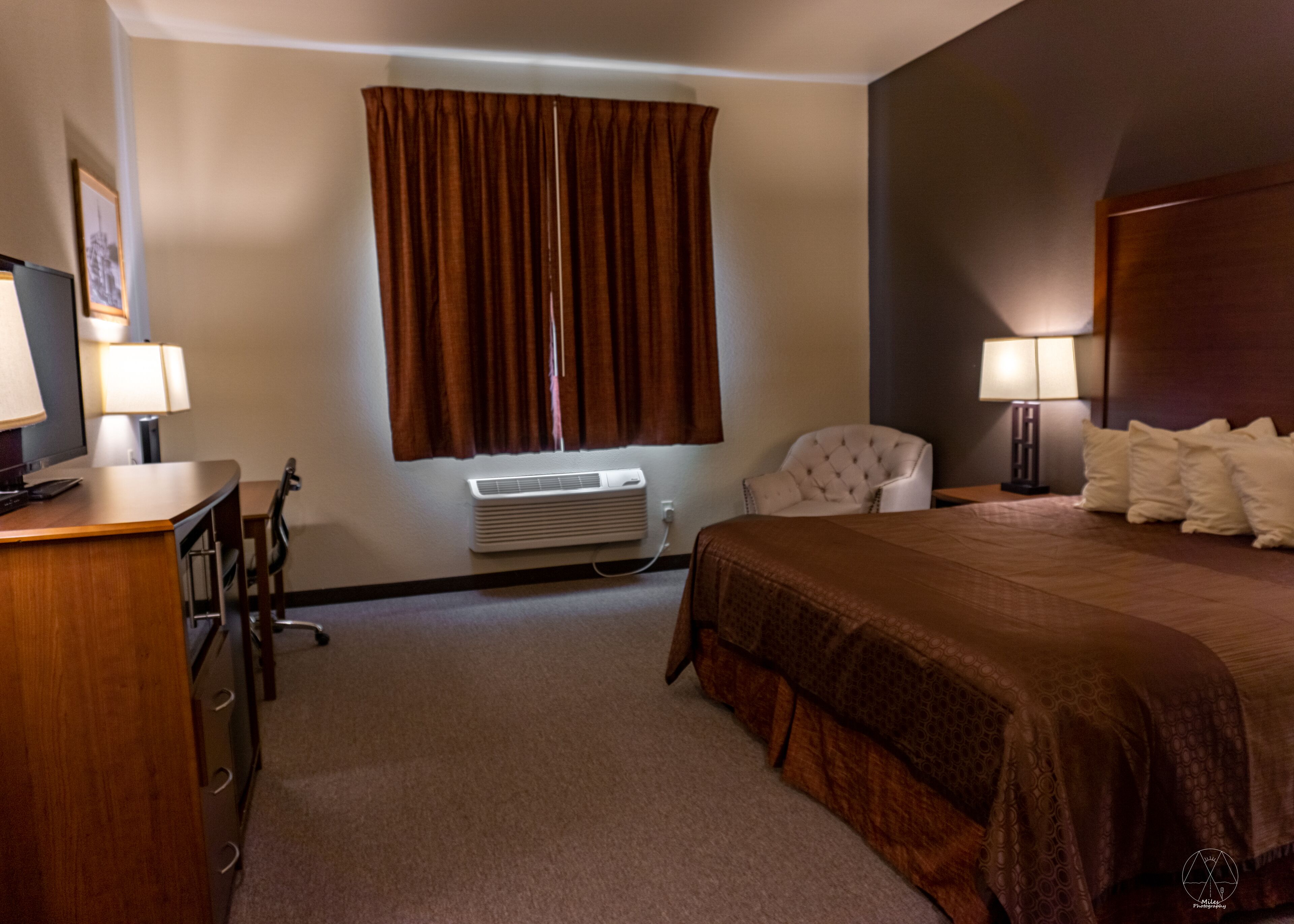 Una habitación de hotel con cama, escritorio, silla, aire acondicionado y ventana con cortinas.