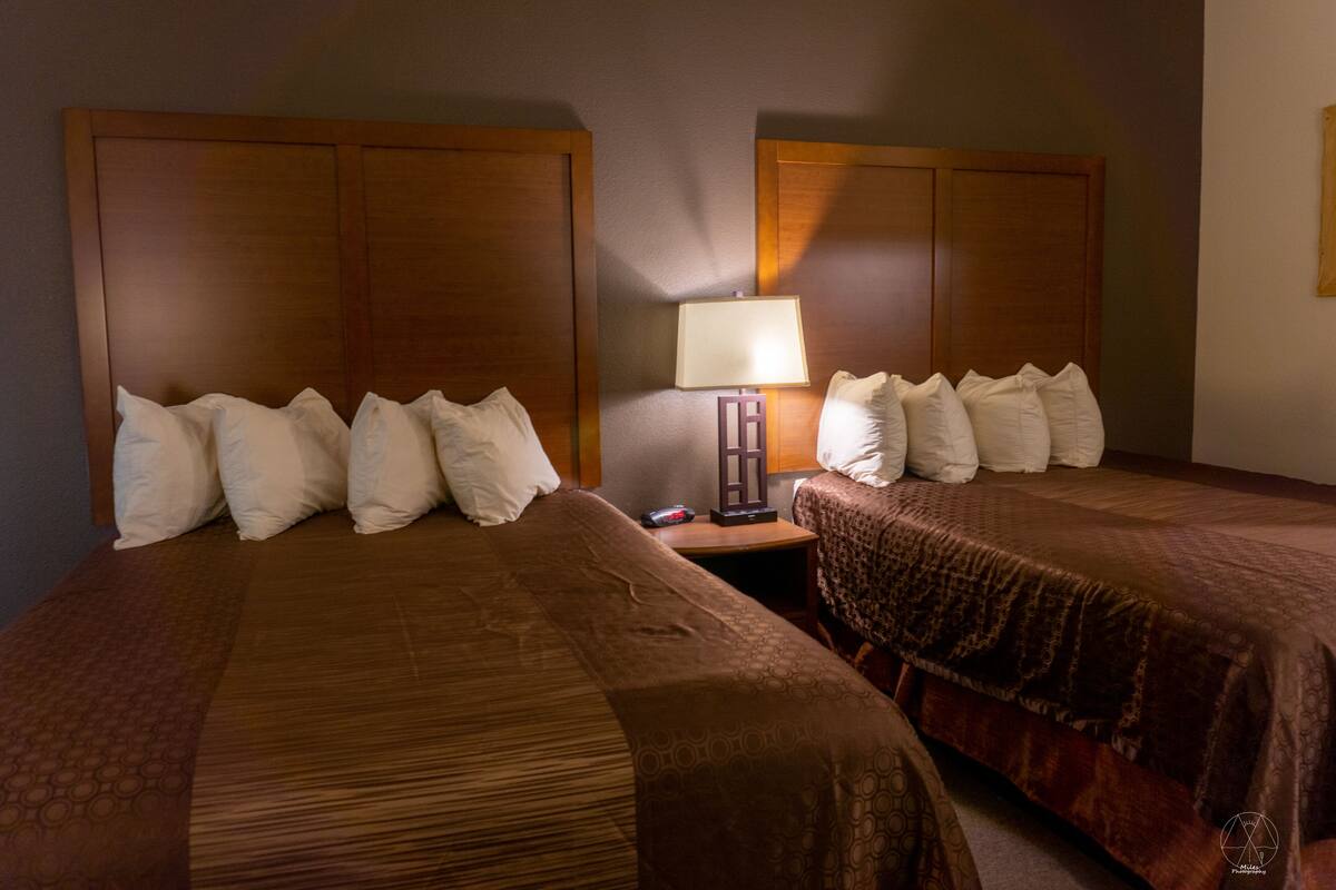 A hotel room with two beds, each with a headboard, a nightstand with a lamp, and several pillows.