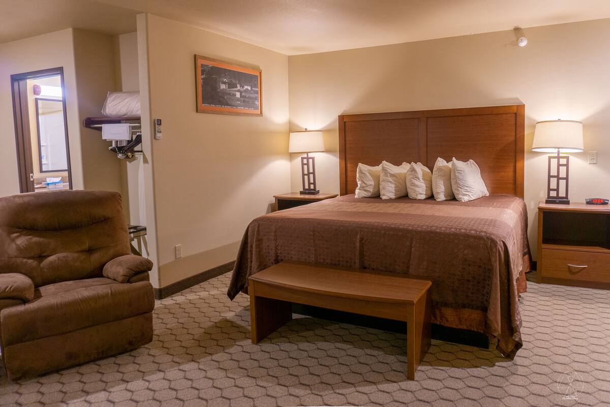 A hotel room with a large bed, a wooden headboard, two bedside lamps, a wooden bench, a chair, and a framed picture on the wall.