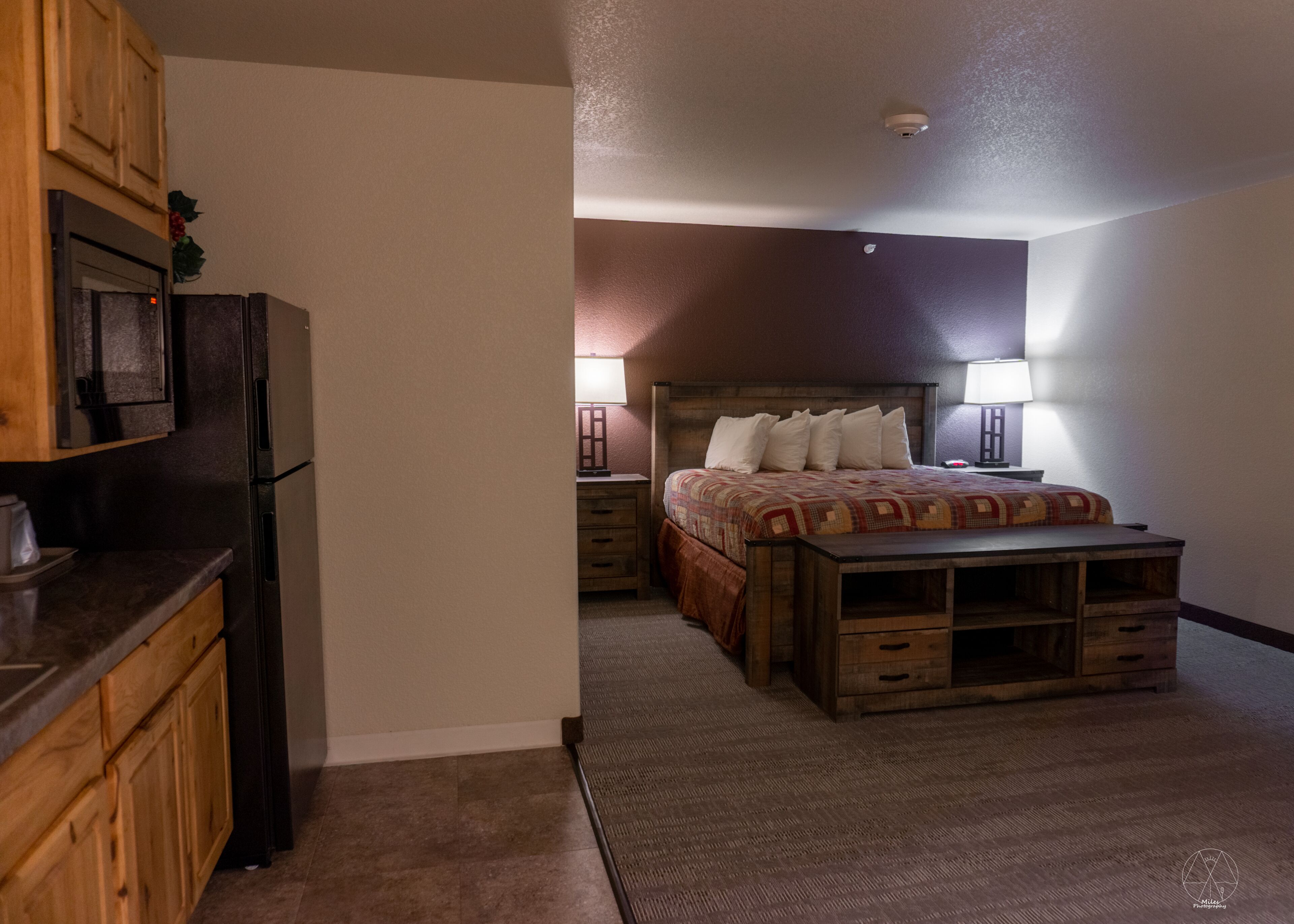 A bedroom with a wooden bed, a nightstand, a lamp, and a refrigerator.