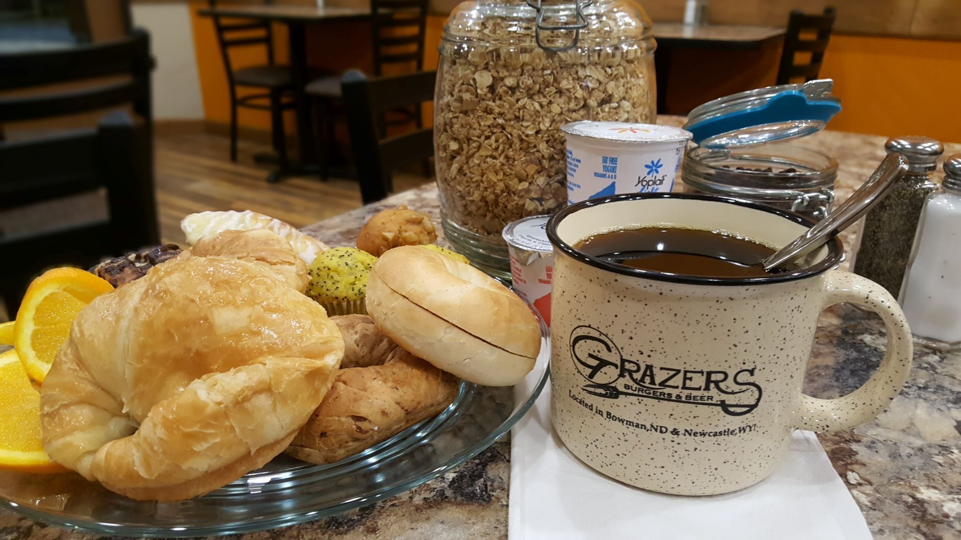 A breakfast setting with pastries, a jar of granola, a mug labeled 'Grazers Burgers & Beer', and a cup of coffee on a marble countertop.