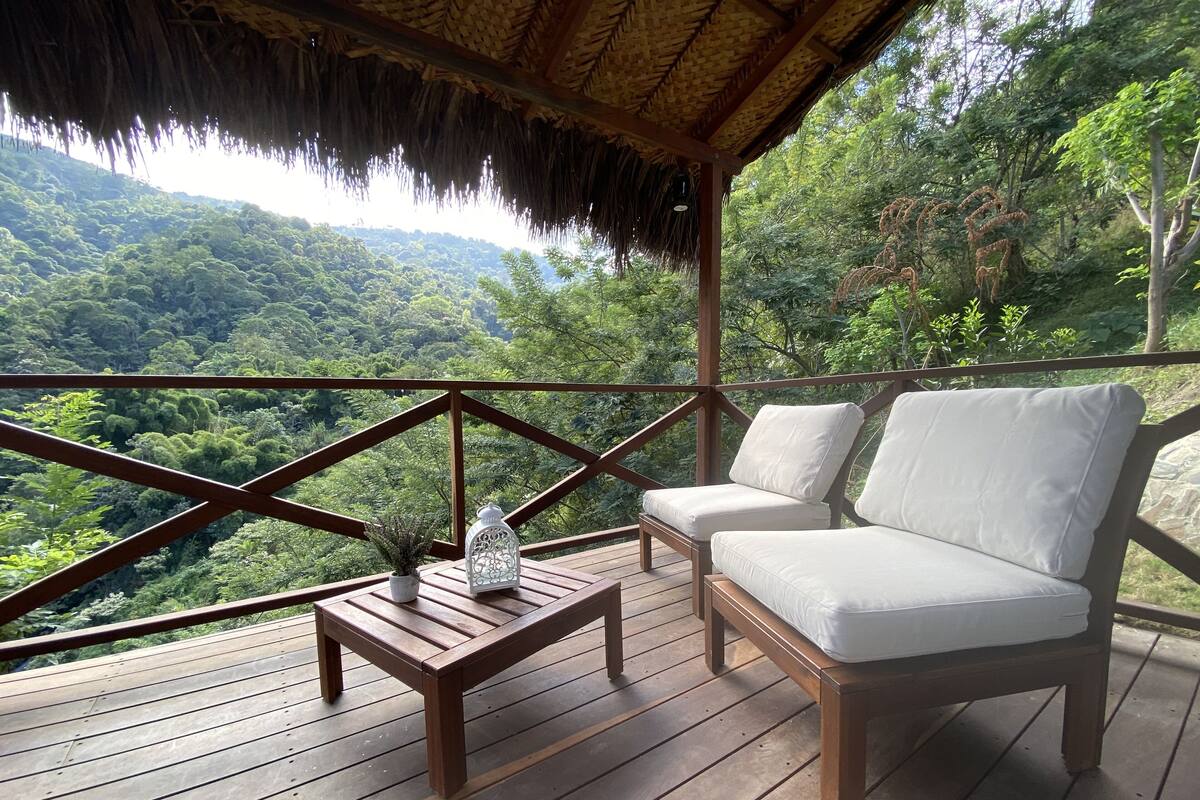 A wooden deck with a thatched roof, a white cushioned sofa, and a small table with a potted plant and a lantern.