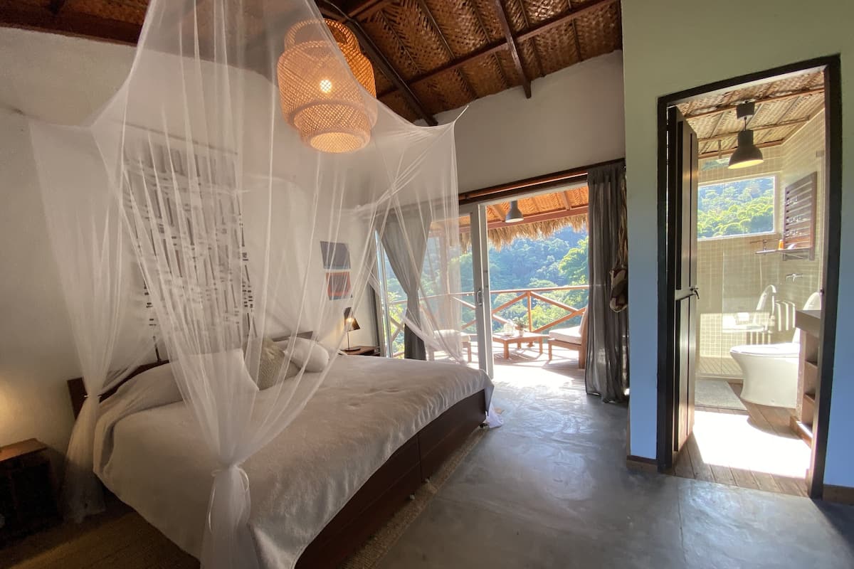 A bedroom with a bed under a mosquito net, a wooden ceiling, and a view of a balcony with a wooden railing.