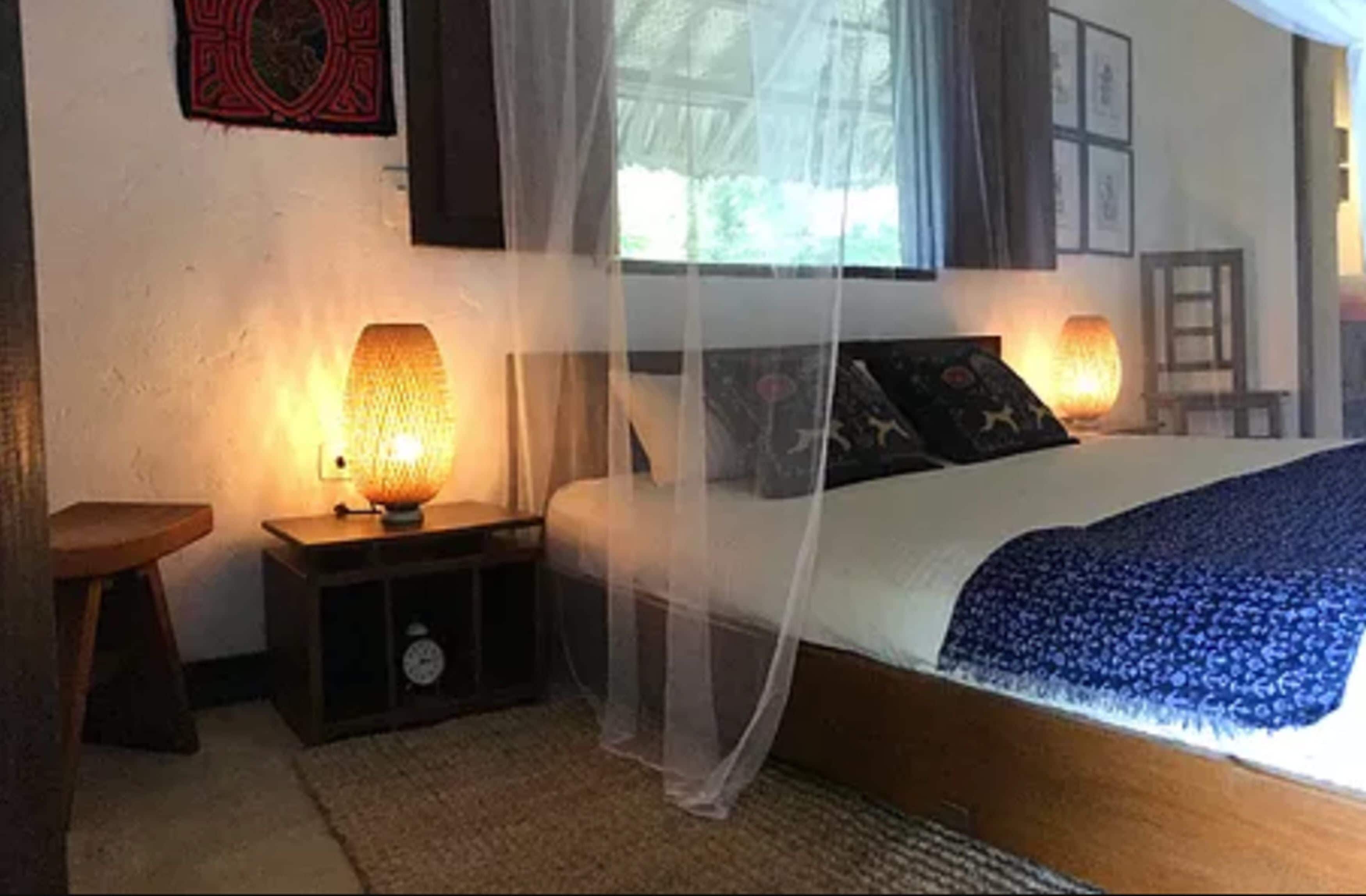 Un dormitorio con una cama cubierta por una mosquitera, dos lámparas de noche, un reloj y un taburete de madera.