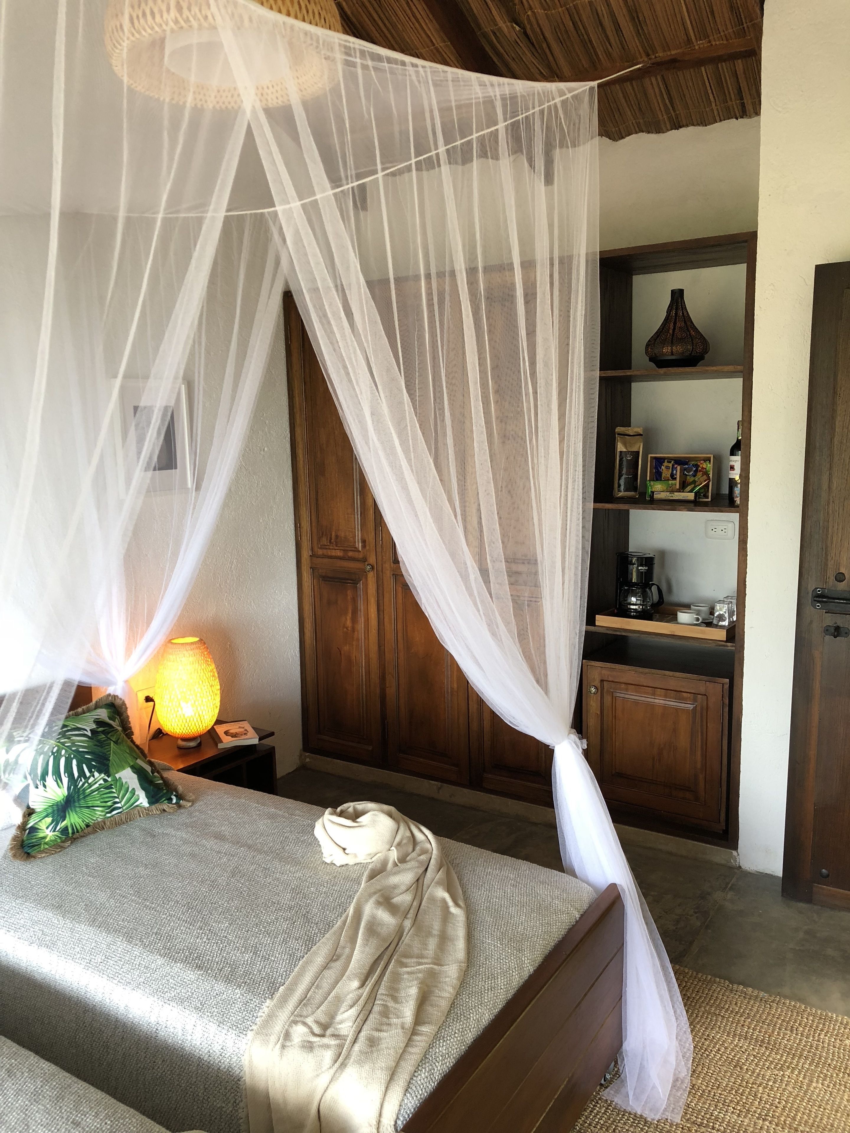 Un dormitorio con una cama bajo una mosquitera, una lámpara de noche y un aparador de madera con estantes.