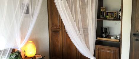 Un dormitorio con una cama bajo una mosquitera, una lámpara de noche y un aparador de madera con estantes.