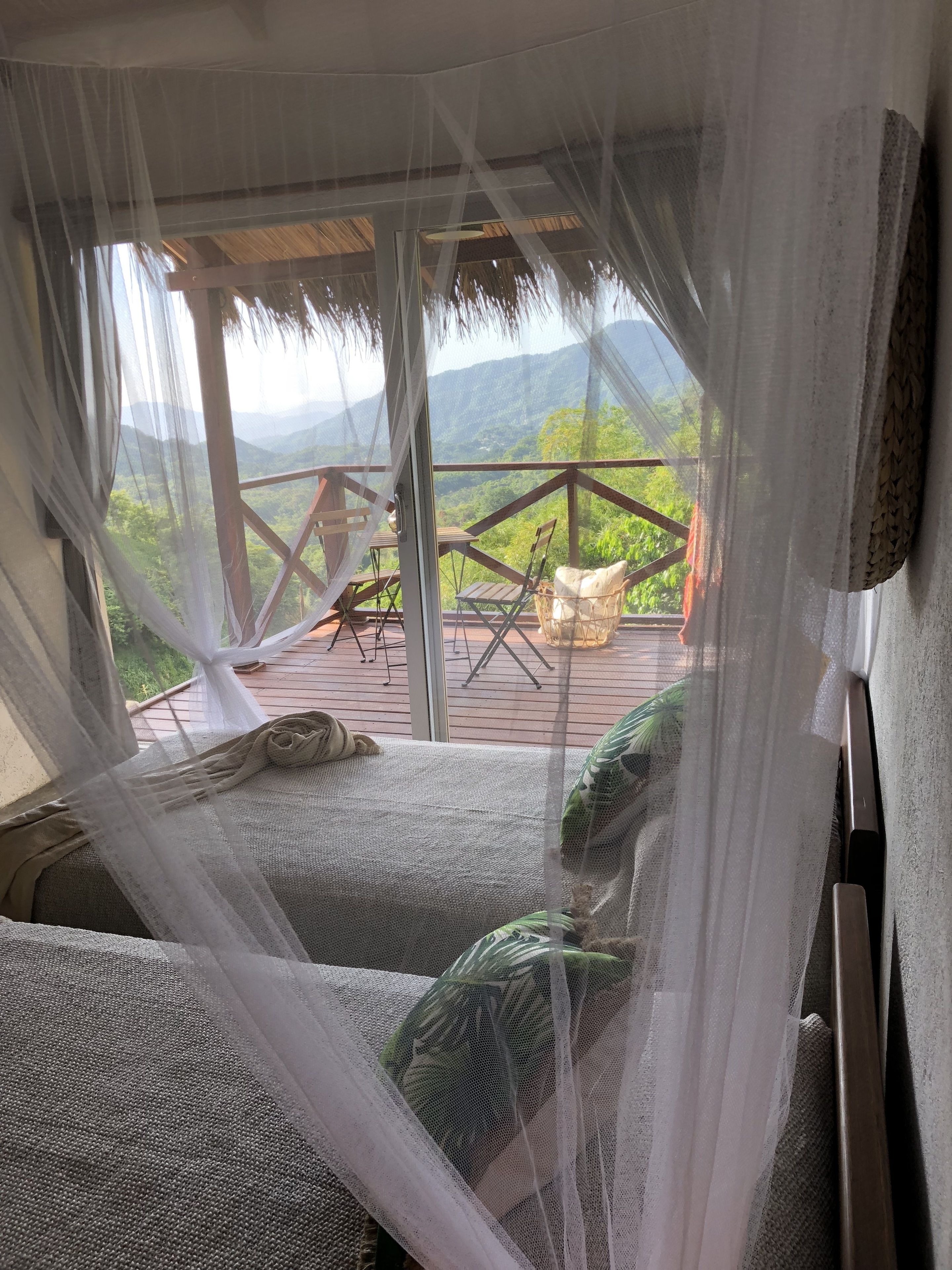 A room with a bed covered by a mosquito net, a wooden deck with chairs, and a view of a mountainous landscape.