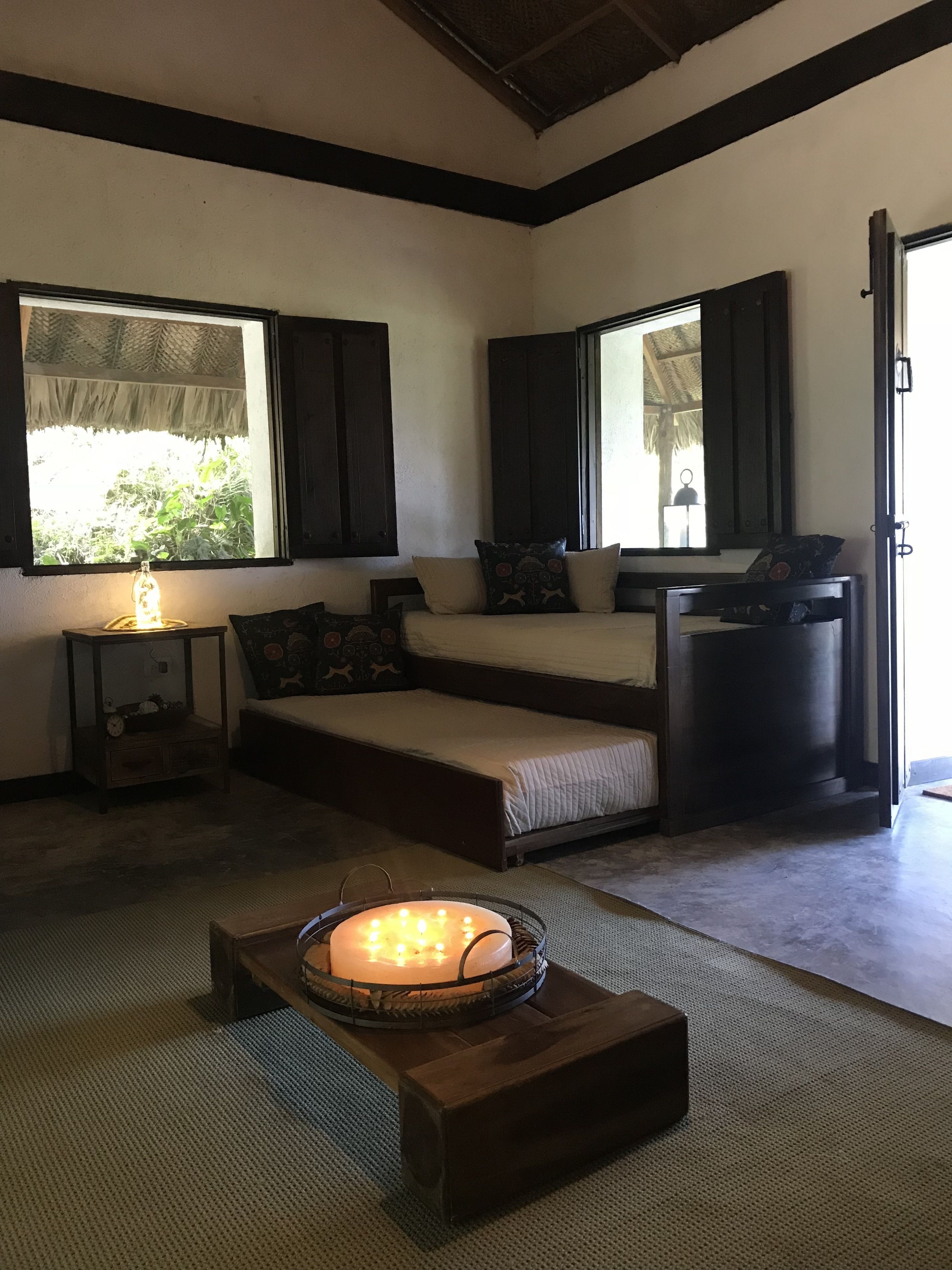 A cozy room with a bed, a bedside table with a lamp, a small table with a candle, and a large window with shutters.