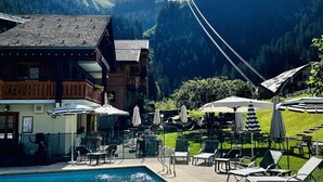 A pool area with lounge chairs and umbrellas, a wooden building with balconies, and a mountainous backdrop. - Hameau des Prodains - Hôtel (Morzine)