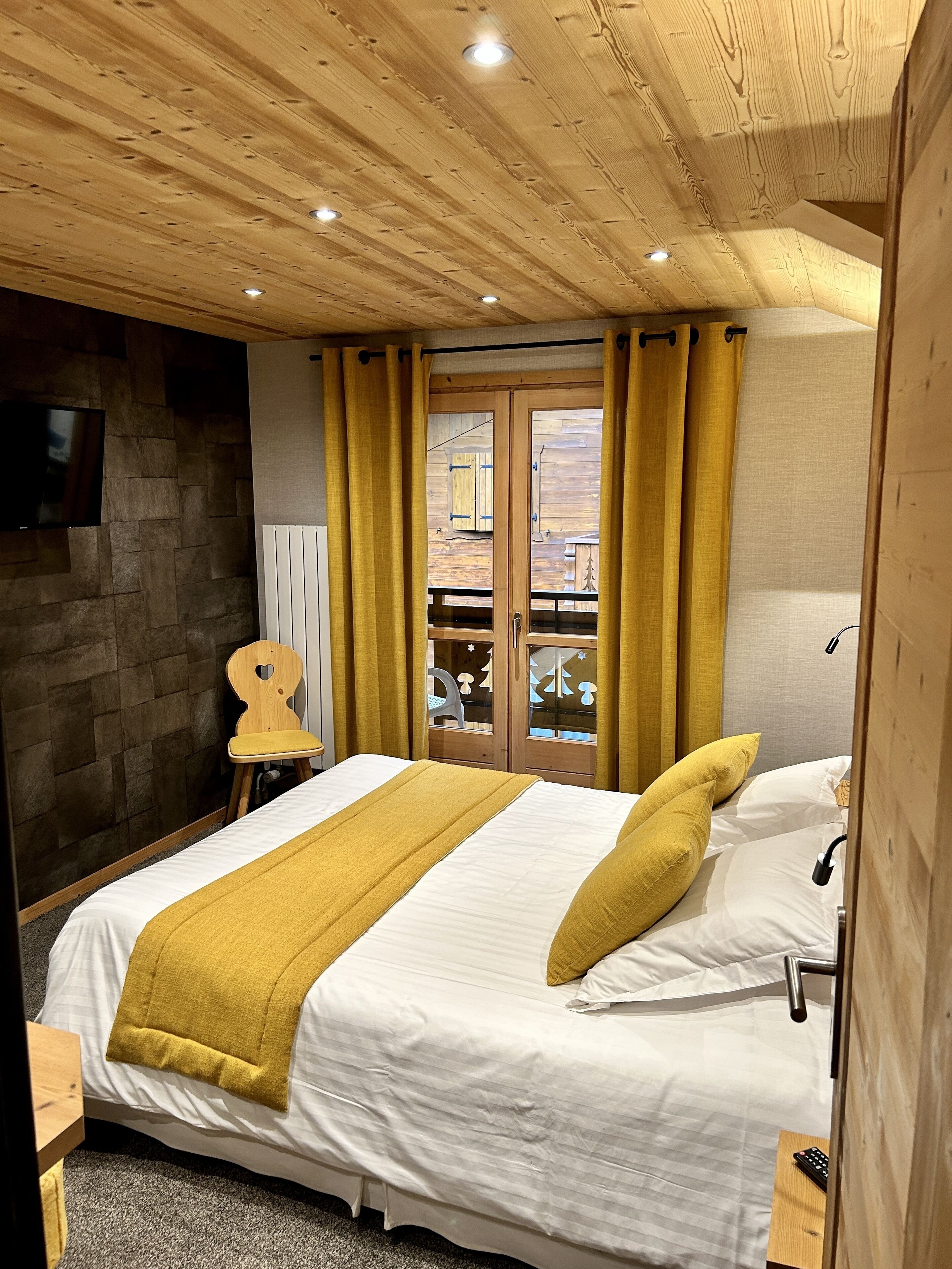 A bedroom with a wooden ceiling, a bed with white bedding and a mustard-colored throw, a wooden chair, and a view through a glass door to an outdoor area.