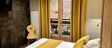 A bedroom with a wooden ceiling, a bed with white bedding and a mustard-colored throw, a wooden chair, and a view through a glass door to an outdoor area.