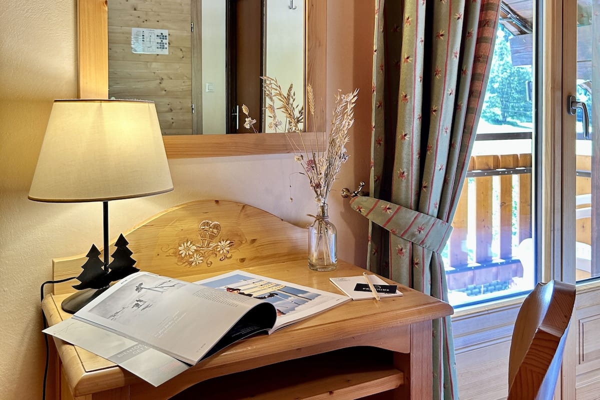 Un bureau en bois, avec une lampe, des papiers et un vase contenant des fleurs séchées. Un miroir reflète une pièce dont le plafond est en bois et qui possède une porte.