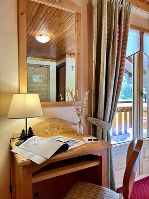 Un bureau en bois, avec une lampe, des papiers et un vase contenant des fleurs séchées. Un miroir reflète une pièce dont le plafond est en bois et qui possède une porte.