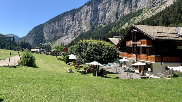 A mountain lodge with outdoor seating and a bar, surrounded by lush greenery and a steep mountain backdrop. - Hameau des Prodains - Hôtel (Morzine)