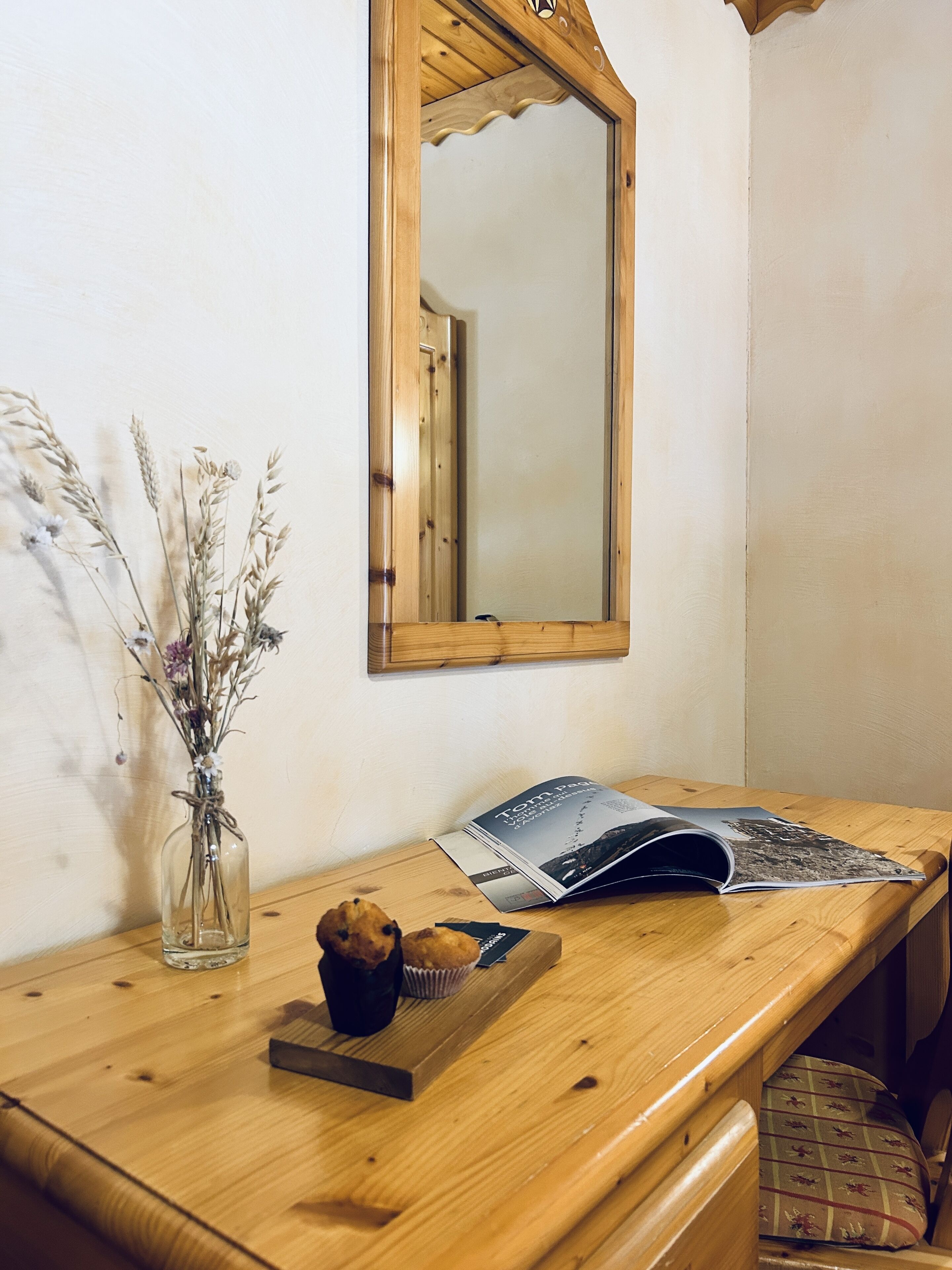 A wooden desk with a vase of dried flowers, a muffin, and a magazine.
