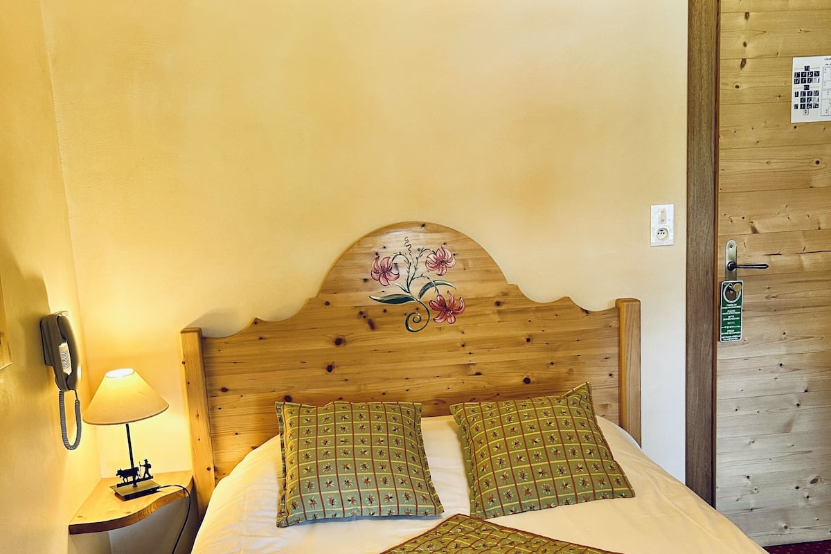 Une chambre d’hôtel avec une tête de lit en bois, deux oreillers à motifs floraux, un lit recouvert d’une courtepointe verte, une lampe de chevet et une porte en bois avec une pancarte.