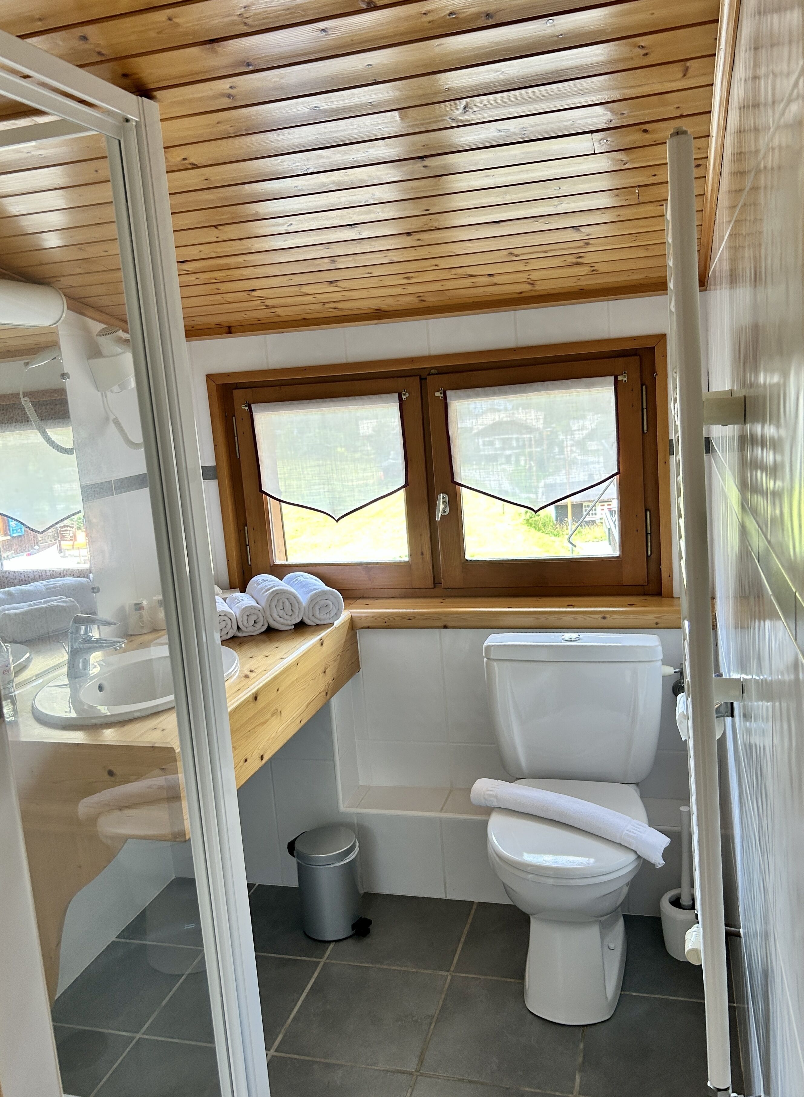 Une petite salle de bain avec un plafond en bois, une fenêtre donnant sur un beau paysage, des toilettes, un lavabo et une petite poubelle.
