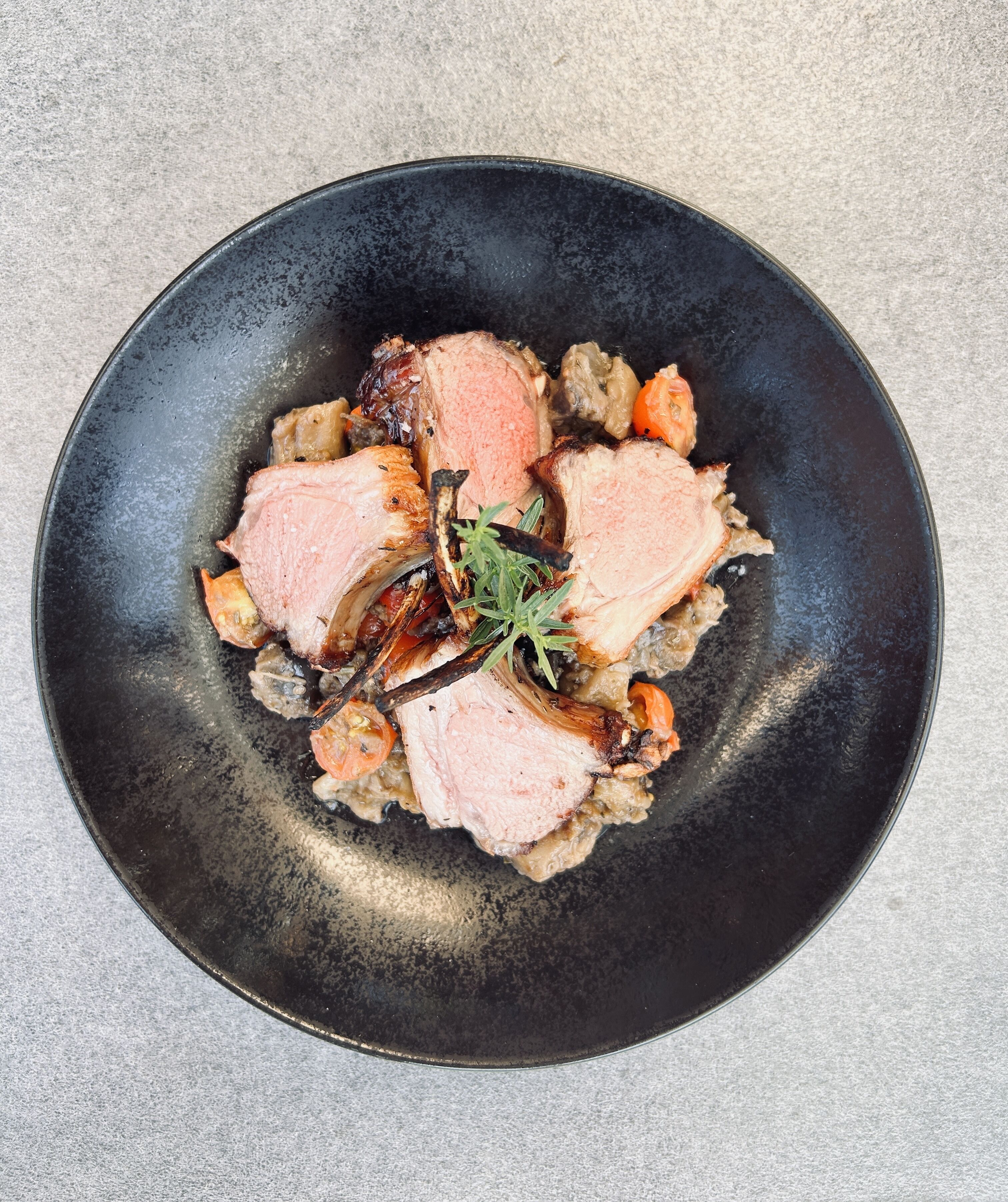 Une assiette noire contenant de la viande tranchée, des légumes et une branche de romarin.