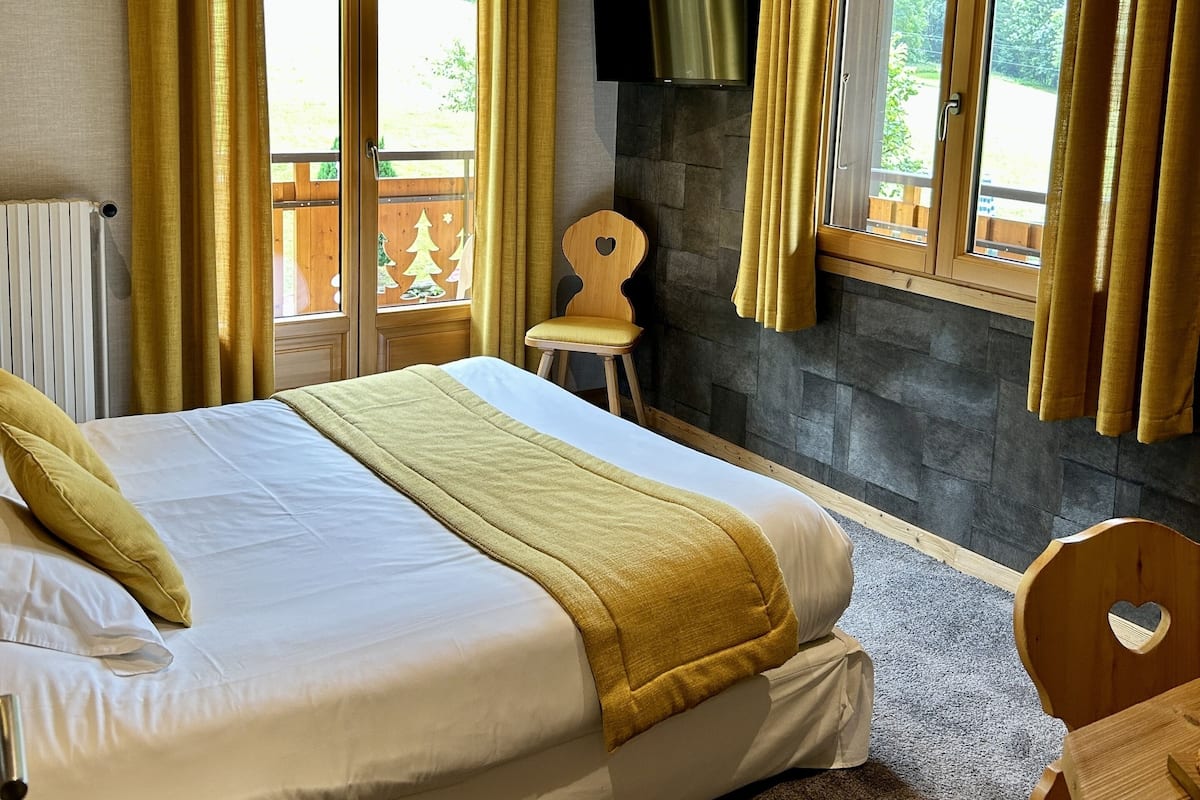 Une chambre avec un grand lit, une tête de lit en bois, un mur en pierre, un téléviseur fixé au mur et une fenêtre avec des rideaux.