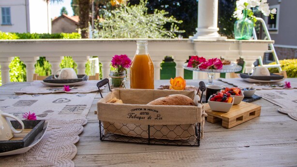 A table set with a bottle, bread, and fruit bowls, with a decorative table runner and a view of a building and greenery in the background. - B&B Villa le Rondini (Castronno)